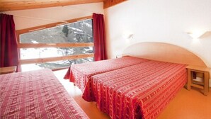 A room with two beds, wooden ceiling beams, a large window with red curtains, and a view of snow-covered trees. - VVF Résidence Plagne 1800 Paradiski (La Plagne-Tarentaise)