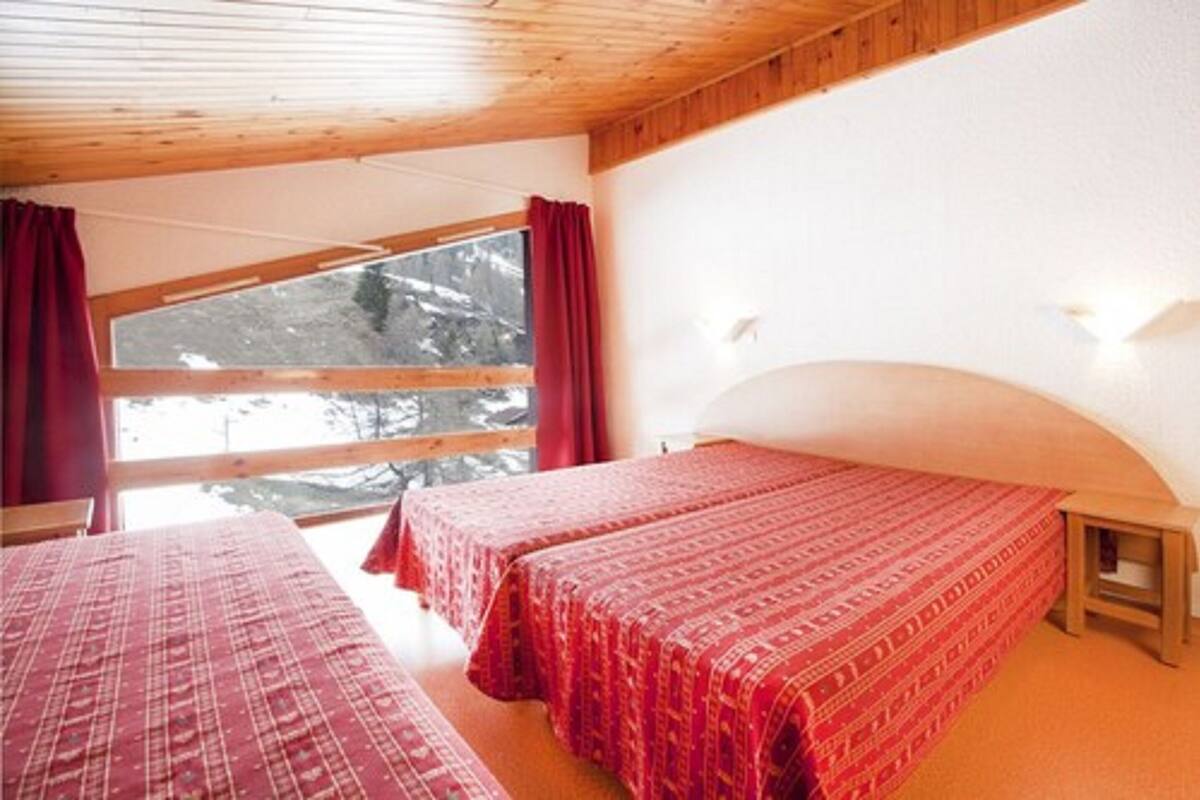 Una habitación con dos camas, vigas de madera en el techo, un ventanal con cortinas rojas y vistas a árboles nevados.