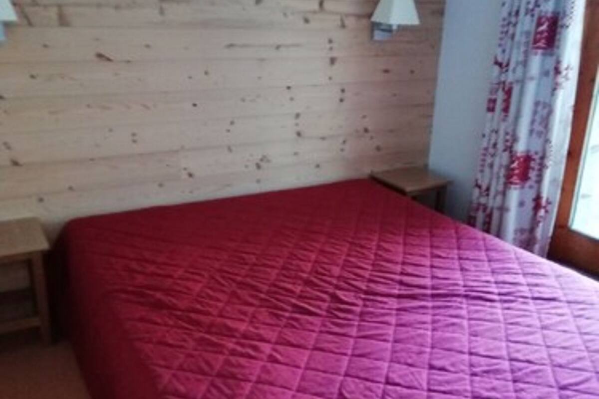 Una habitación con pared de madera, colcha roja, dos mesitas de noche y una ventana con cortinas florales.