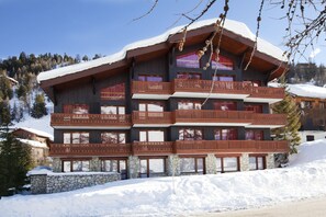 A multi-story chalet with wooden balconies, stone foundation, and a snow-covered roof. - VVF Résidence Plagne 1800 Paradiski (La Plagne-Tarentaise)