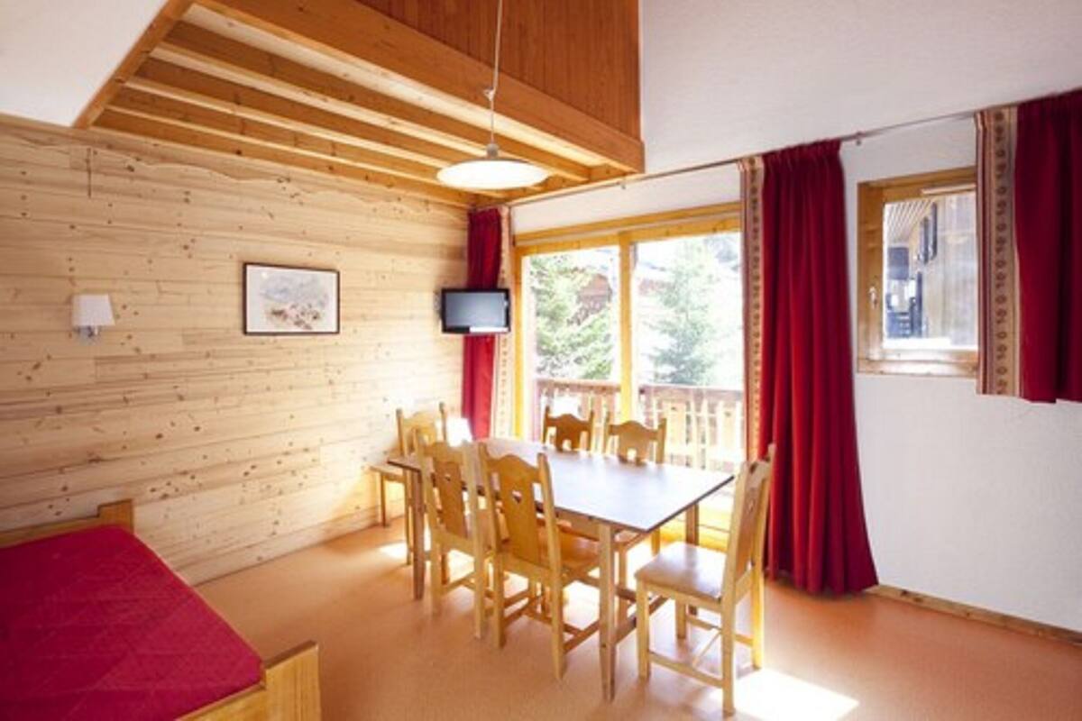 Una habitación con techo de madera, una mesa de comedor con sillas, un televisor montado en la pared y una ventana con cortinas rojas.