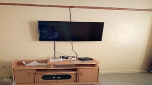 Ein Zimmer mit einem an der Wand befestigten Fernseher auf einem Holzständer, einem kleinen Tisch mit Papieren und einem Glastisch im Vordergrund.