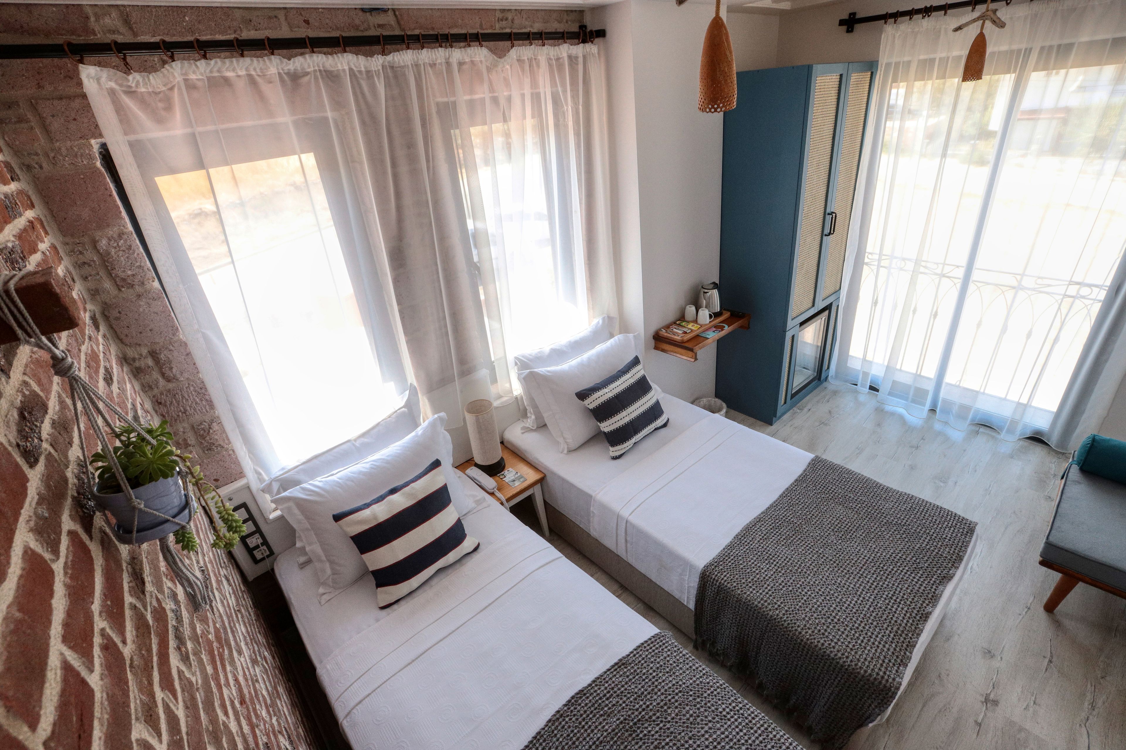 Une chambre avec deux lits, un mur en briques, une fenêtre avec des rideaux légers, une petite table avec une lampe et une plante suspendue.
