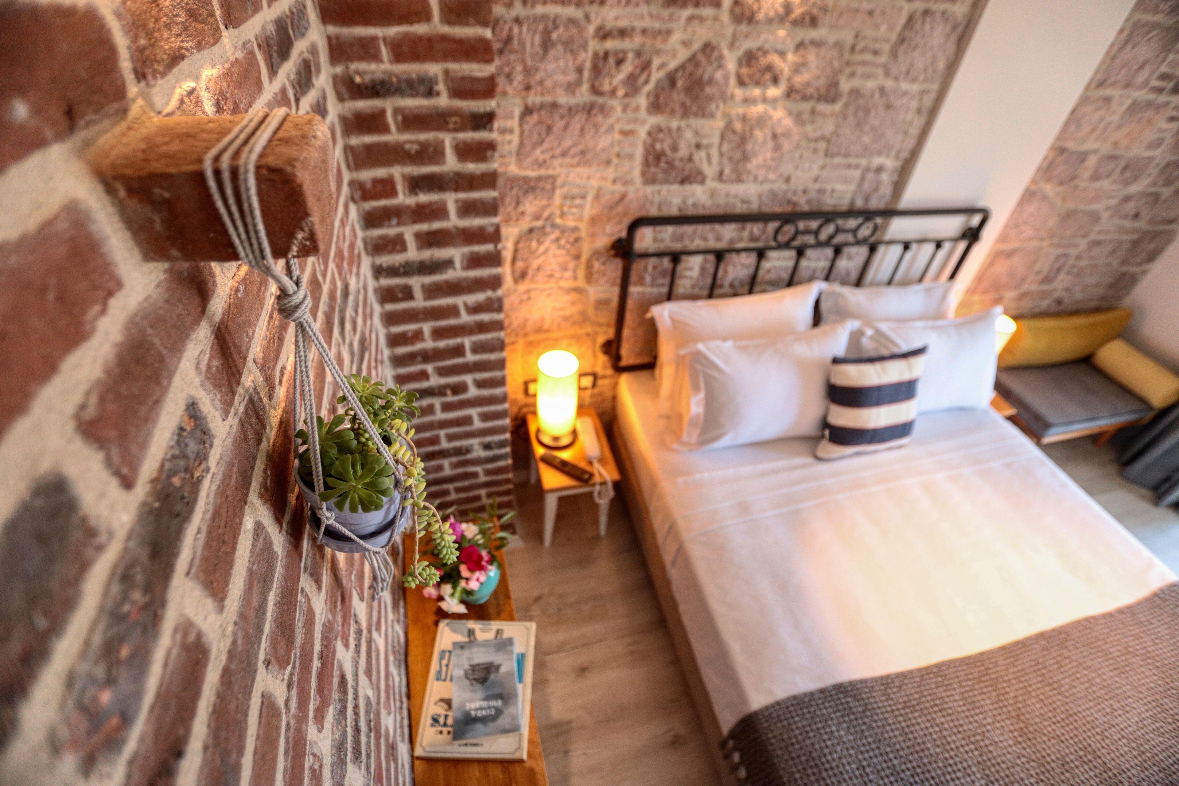 A cozy room with a brick wall, a bed with pillows, a bedside table with a lamp, and a small plant.