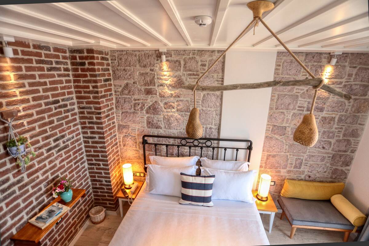 Ein gemütliches Schlafzimmer mit Backsteinwand, einem Bett mit weißen Leinentextilien, einer Hängelampe und einer kleinen Sitzecke mit Kissen.