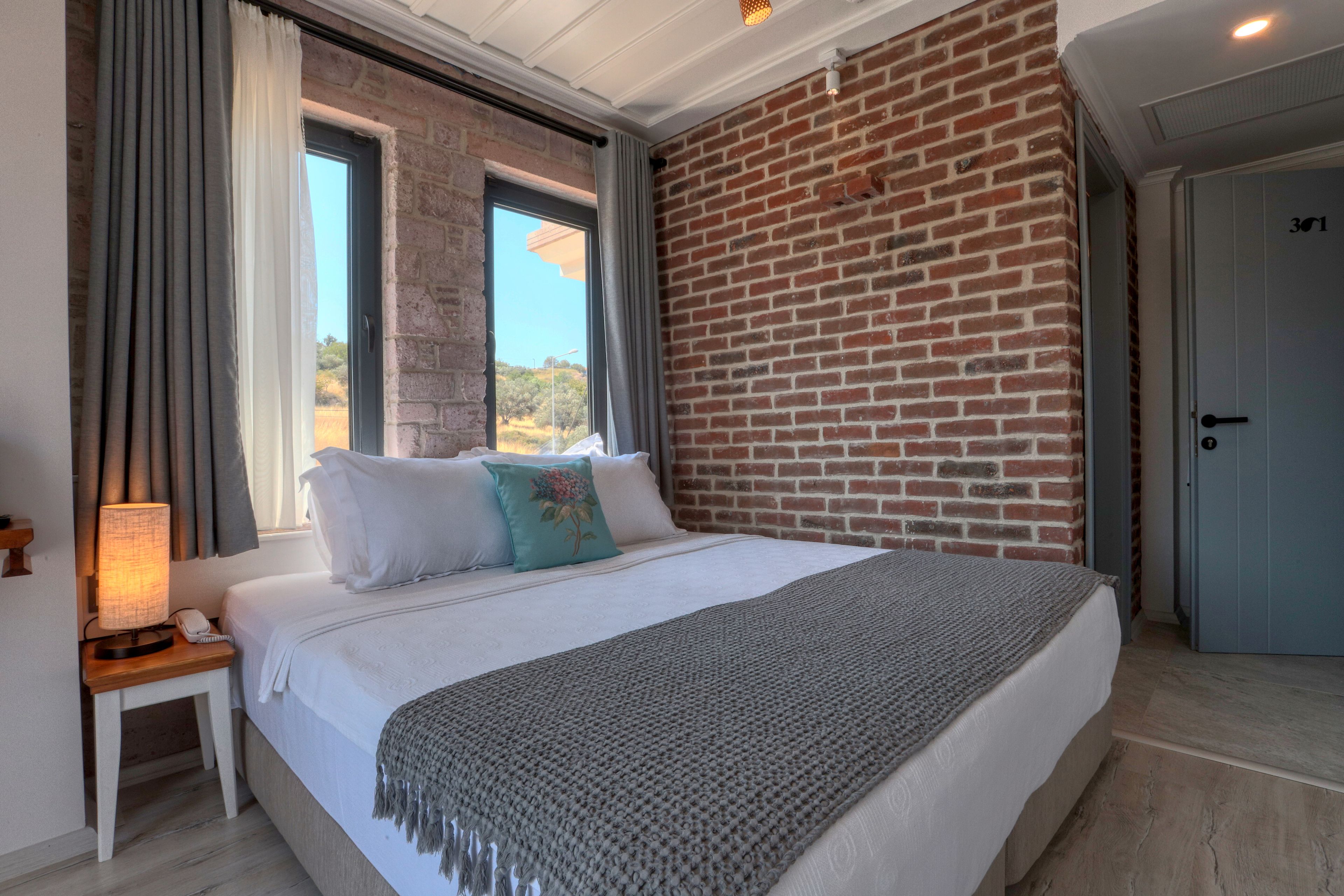 Une chambre à coucher avec un grand lit, une table de chevet avec une lampe et une vue sur l’extérieur par une fenêtre.