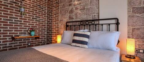 A bedroom with a brick wall, a bed with white bedding, a striped pillow, a small shelf with books and a vase, and a bedside lamp.