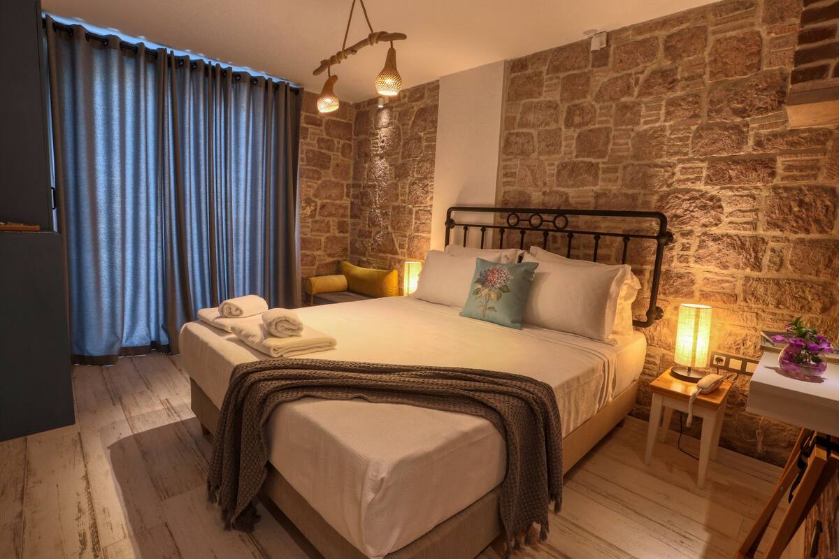 Ein Schlafzimmer mit Steinwand, einem Bett mit Kissen, einem Nachttisch mit Lampe und einem Fenster mit Vorhängen.
