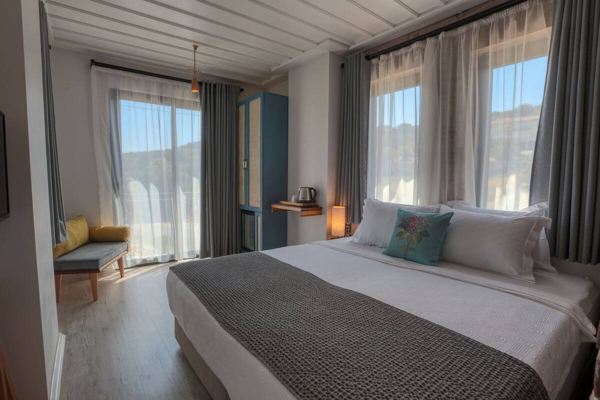 Ein Hotelzimmer mit einem großen Bett, einem Nachttisch mit Lampe, einem Sessel und Blick ins Freie.
