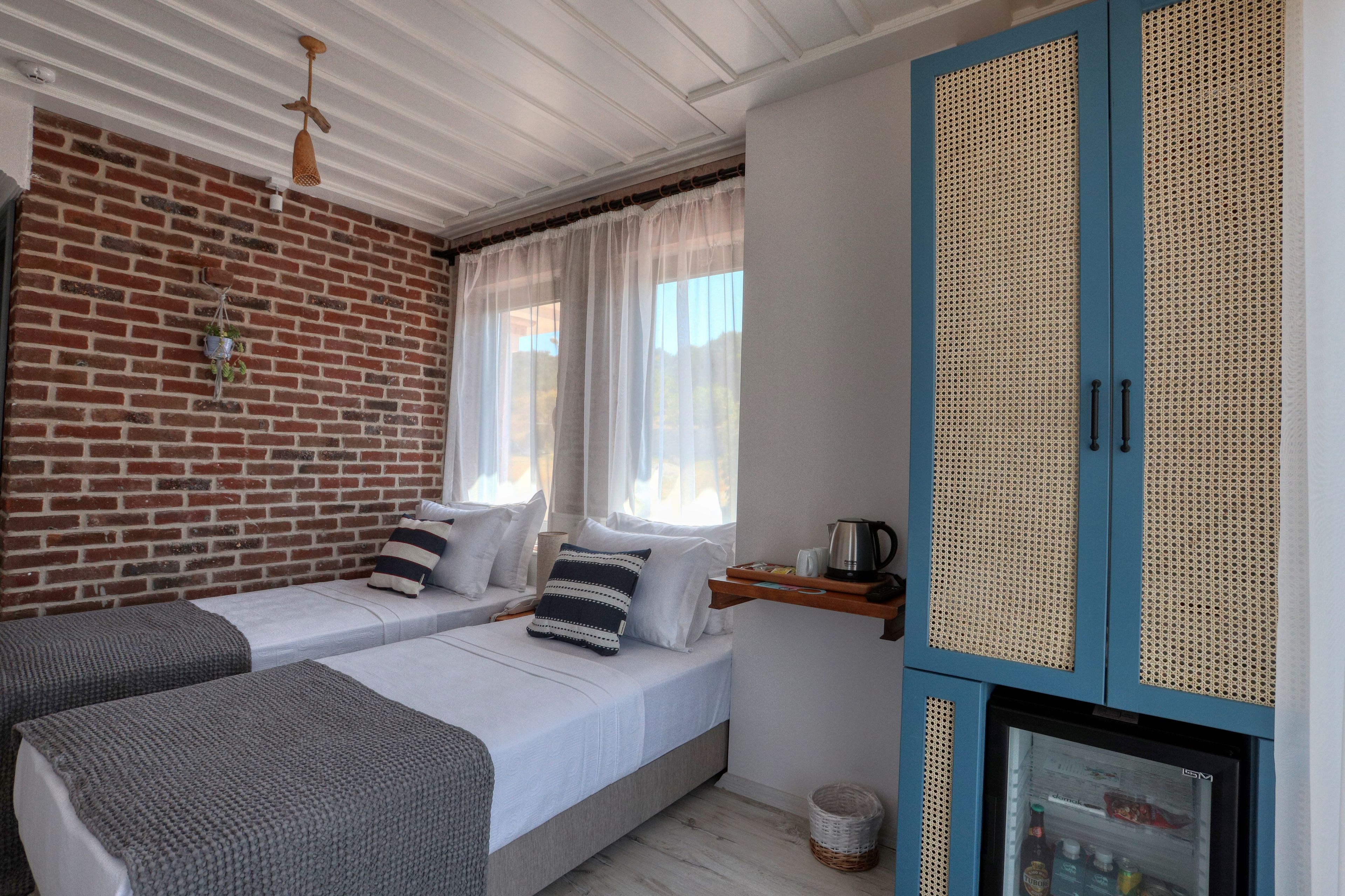 Quarto com duas camas, parede de tijolos, janela com cortinas leves, armário azul e uma geladeira pequena.