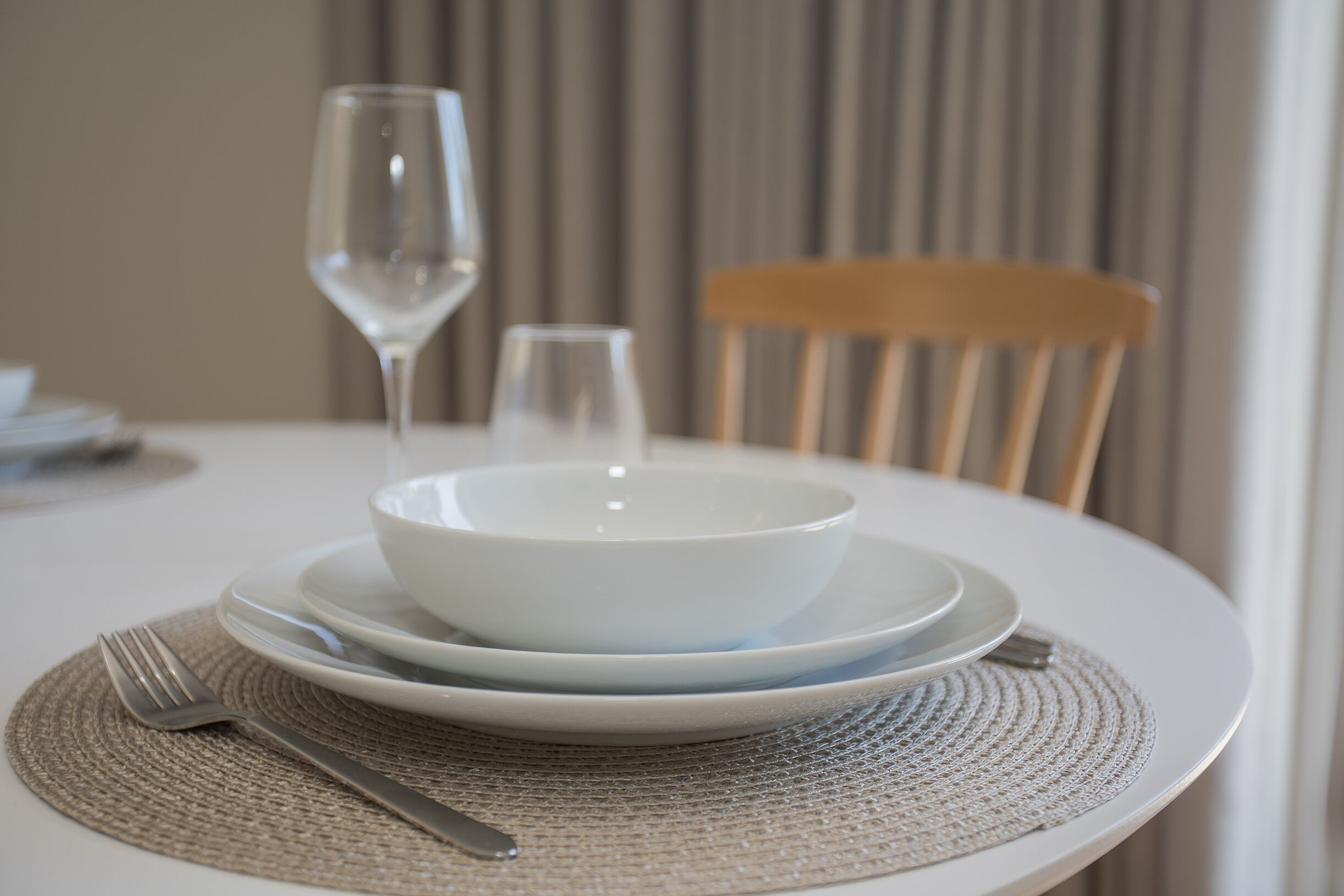 A dining table with a white tablecloth, a woven placemat, a bowl, a wine glass, and a chair.