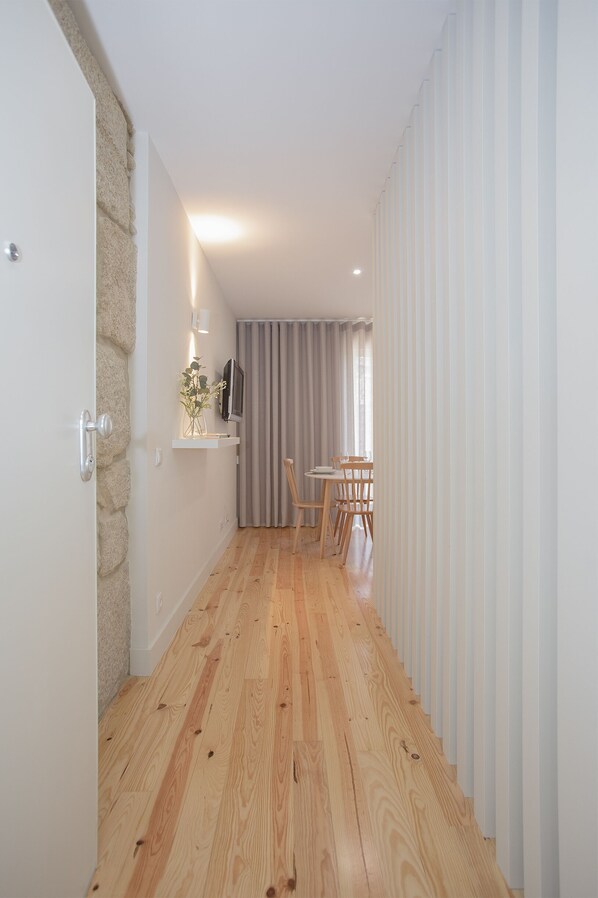 A narrow hallway with wooden flooring, a stone accent wall, a small table with chairs, and a television on a shelf. - Liiiving in Porto - Charming Downtown Delight I (Porto)