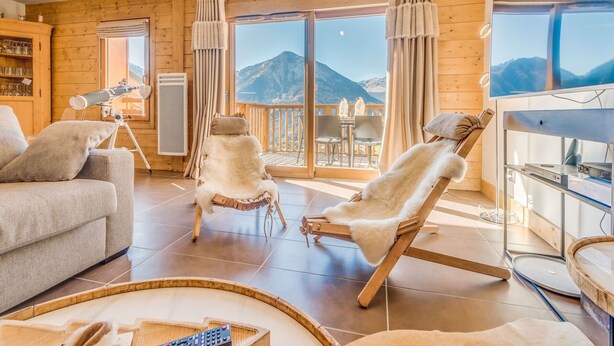 Ein gemütliches Wohnzimmer mit einer Couch, zwei Fellstühlen, einem Teleskop und einem Balkon mit Bergblick.