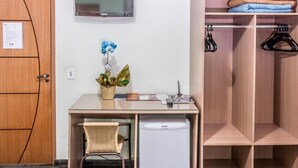 Una habitación compacta con un escritorio de madera, un refrigerador pequeño, una silla, un televisor fijado a la pared y un armario con ropa colgada.