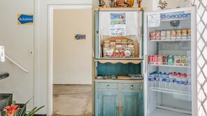Una kitchenette con un refrigerador lleno de bebidas, un estante pequeño con botanas, y una puerta con un letrero que indica el número de habitación.