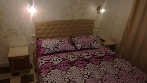 A bedroom with a tufted headboard, a patterned bedspread, two bedside tables, and wall sconces. - Hotel Djhaltia (Ténès)