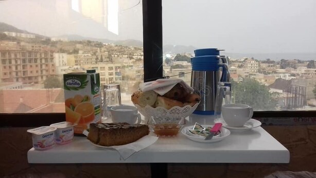 A breakfast table with a carton of orange juice, a thermos, a basket of bread, a plate with a pastry, and a cup of coffee. - Hotel Djhaltia (Ténès)