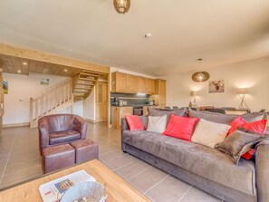 Ein geräumiger Wohnbereich mit einer Couch, einem Ledersessel und einem Holztisch. Im Hintergrund ist eine Küche erkennbar, mit Holzschränken und einer Treppe, die zu einer weiteren Ebene führt.