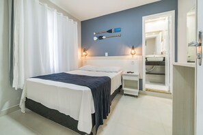 A bedroom with a bed, a nightstand, a mirror, and a bathroom visible through an open door. - Residencial Bombinhas (Bombinhas)