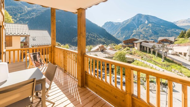 Une terrasse en bois avec une table et des chaises, offrant une vue sur un paysage montagneux parsemé de bâtiments et sous un ciel dégagé.
