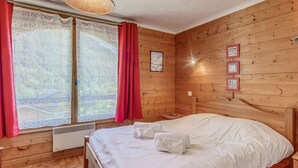 Ein Zimmer in einer Holzhütte mit einem Bett, einem Fenster mit Vorhängen, einem an der Wand hängenden Bild und einer Deckenleuchte.