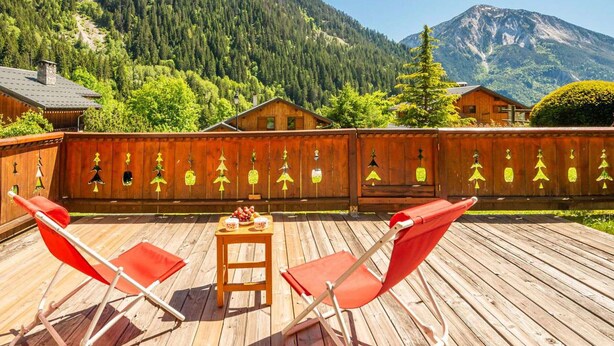 Eine Holzdar Terrasse mit roten Stühlen und einem Tisch, mit Blick auf eine bewaldete Bergkette.