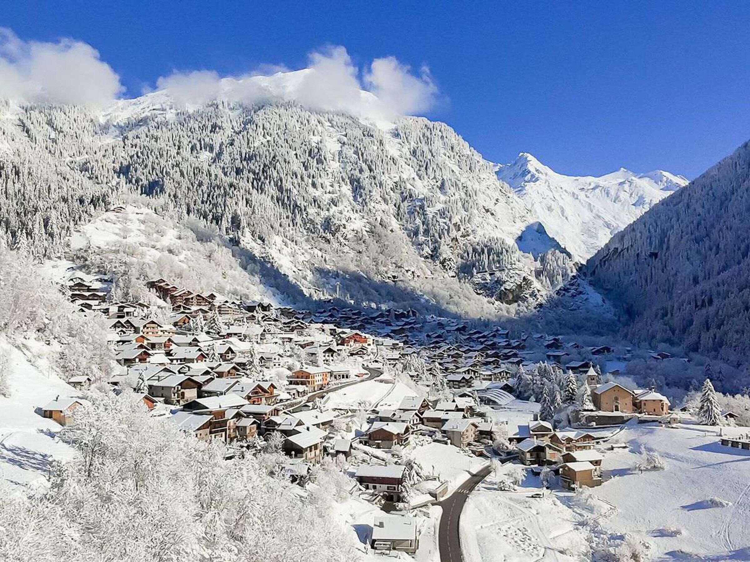 A snow-covered village with traditional wooden houses, a winding road, and mountains in the background.
