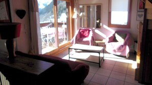 Ein gemütliches Wohnzimmer mit einer Couch, einem Couchtisch und einem Sessel. Ein großes Fenster bietet Blick auf die Berge.