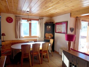 A cozy dining area with wooden furniture, a round table, and chairs. There is a window with curtains, a wall clock, and a cabinet with a mirror.