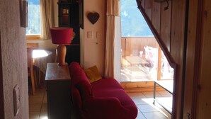 Ein gemütliches Zimmer mit Holzbalkendecke, einem roten Sessel, einem Tisch mit Lampe und Blick auf einen Balkon mit Sitzmöbeln.
