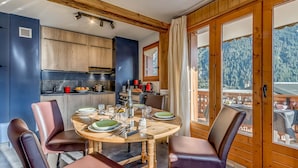 A dining area with a wooden table set for four, surrounded by leather chairs. A kitchen with wooden cabinets and a view of a mountainous landscape is visible through a glass door. - Champagny en Vanoise - Renovated 2-room, 6 pers, balcony, WIFI (Champagny-en-Vanoise)