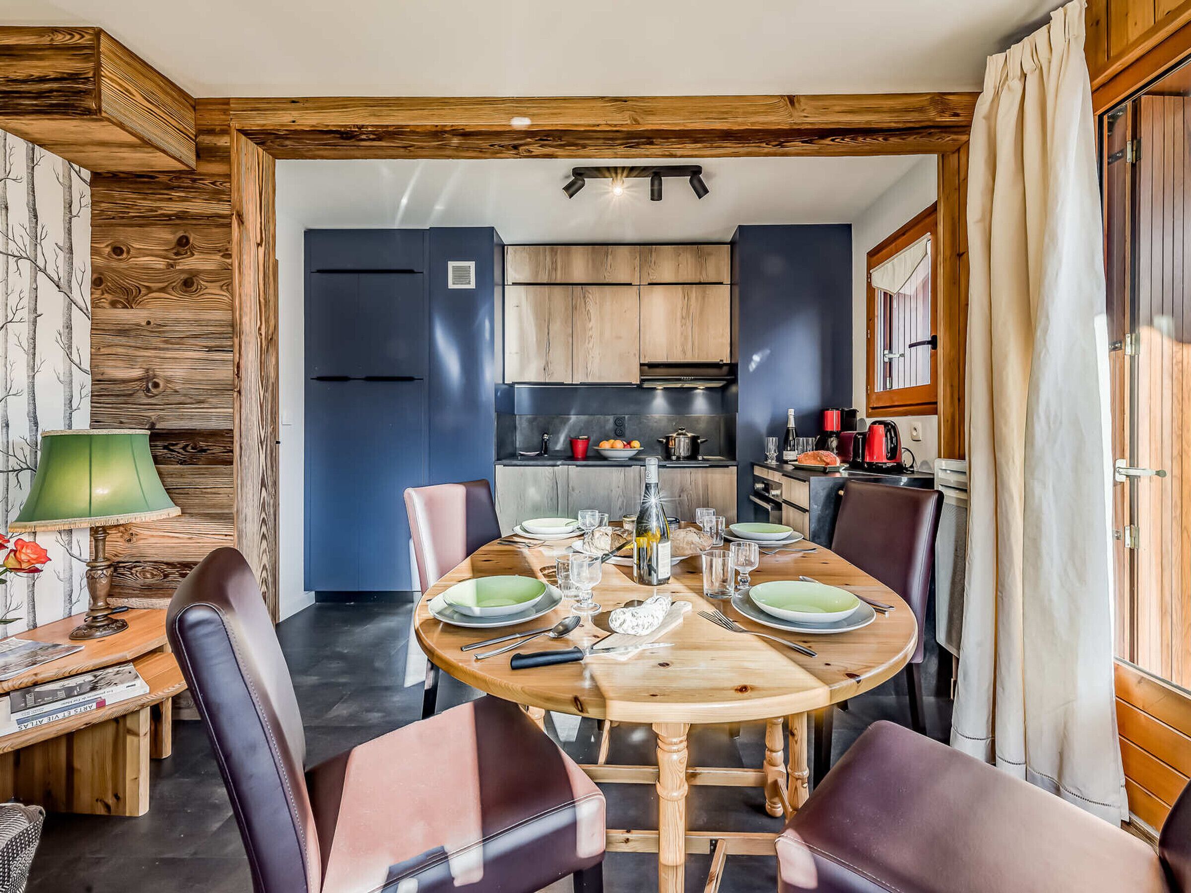 A dining area with a wooden table set for four, surrounded by leather chairs. A kitchen is visible in the background with wooden cabinets and modern appliances.