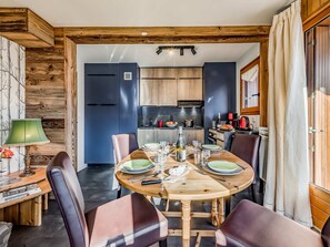 Uma sala de jantar com uma mesa de madeira posta para quatro pessoas, rodeada por cadeiras de couro. Uma cozinha visível ao fundo, com armários de madeira e eletrodomésticos modernos.