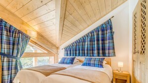 A cozy bedroom with two beds, plaid curtains, a wooden ceiling, and a nightstand with a lamp.