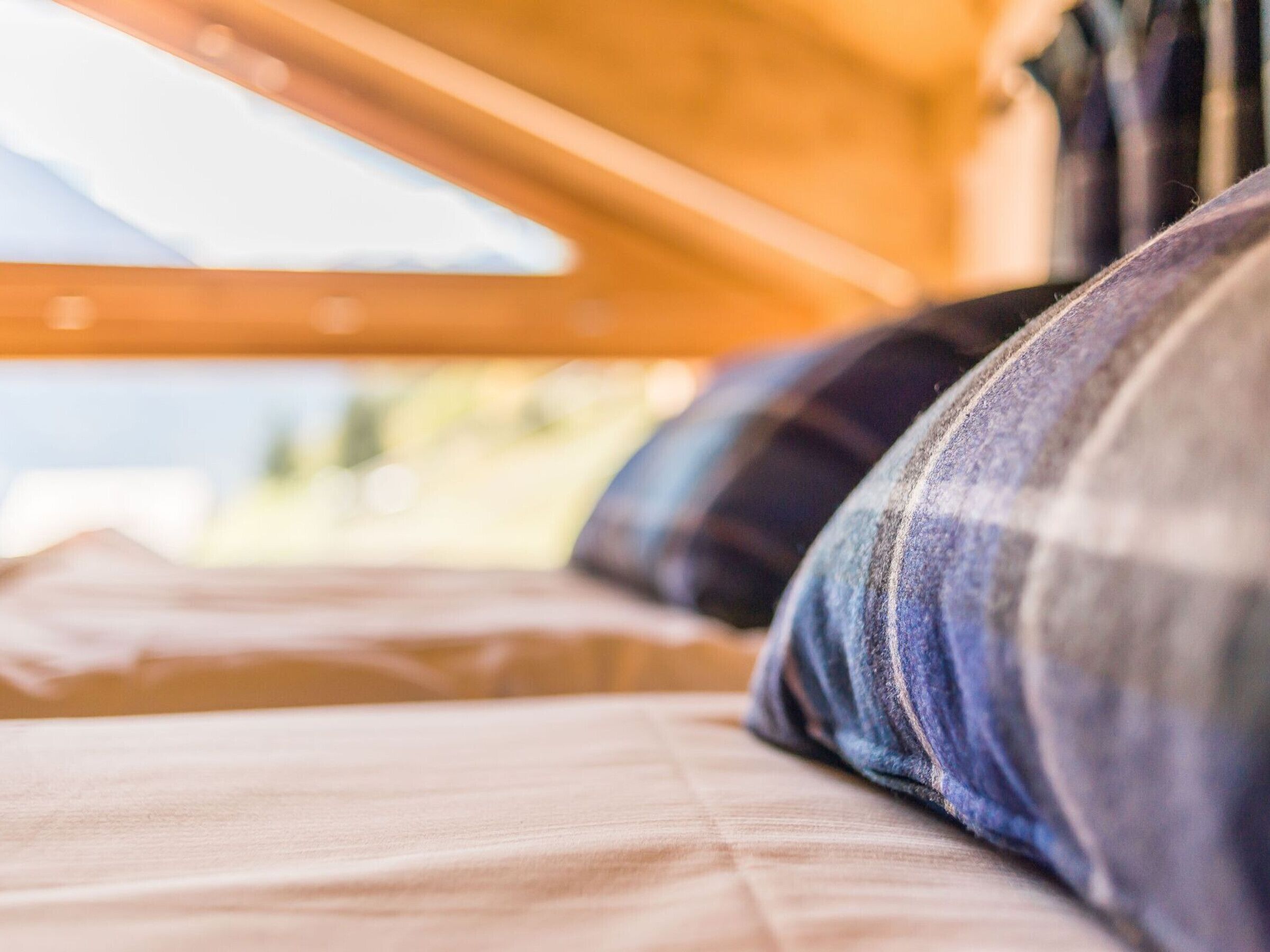 Un gros plan d’un lit avec une couverture à carreaux et des oreillers, une tête de lit en bois et une fenêtre donnant sur un paysage verdoyant.