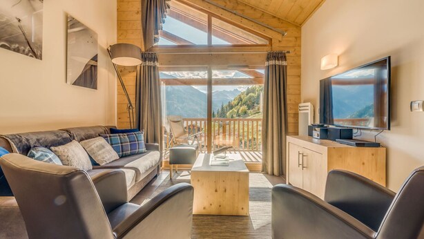 A cozy living room with a sofa, chairs, a coffee table, and a TV. There is a balcony with a view of mountains.