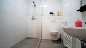 A small, white-tiled bathroom with a shower, toilet, and sink. There are toiletries on the sink and a plant on the shelf. - Ferienwohnung Maye Oberhausen (Oberhausen)