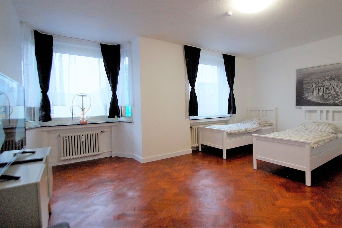Quarto com duas camas de solteiro, um radiador, janela com cortinas pretas e um quadro na parede.