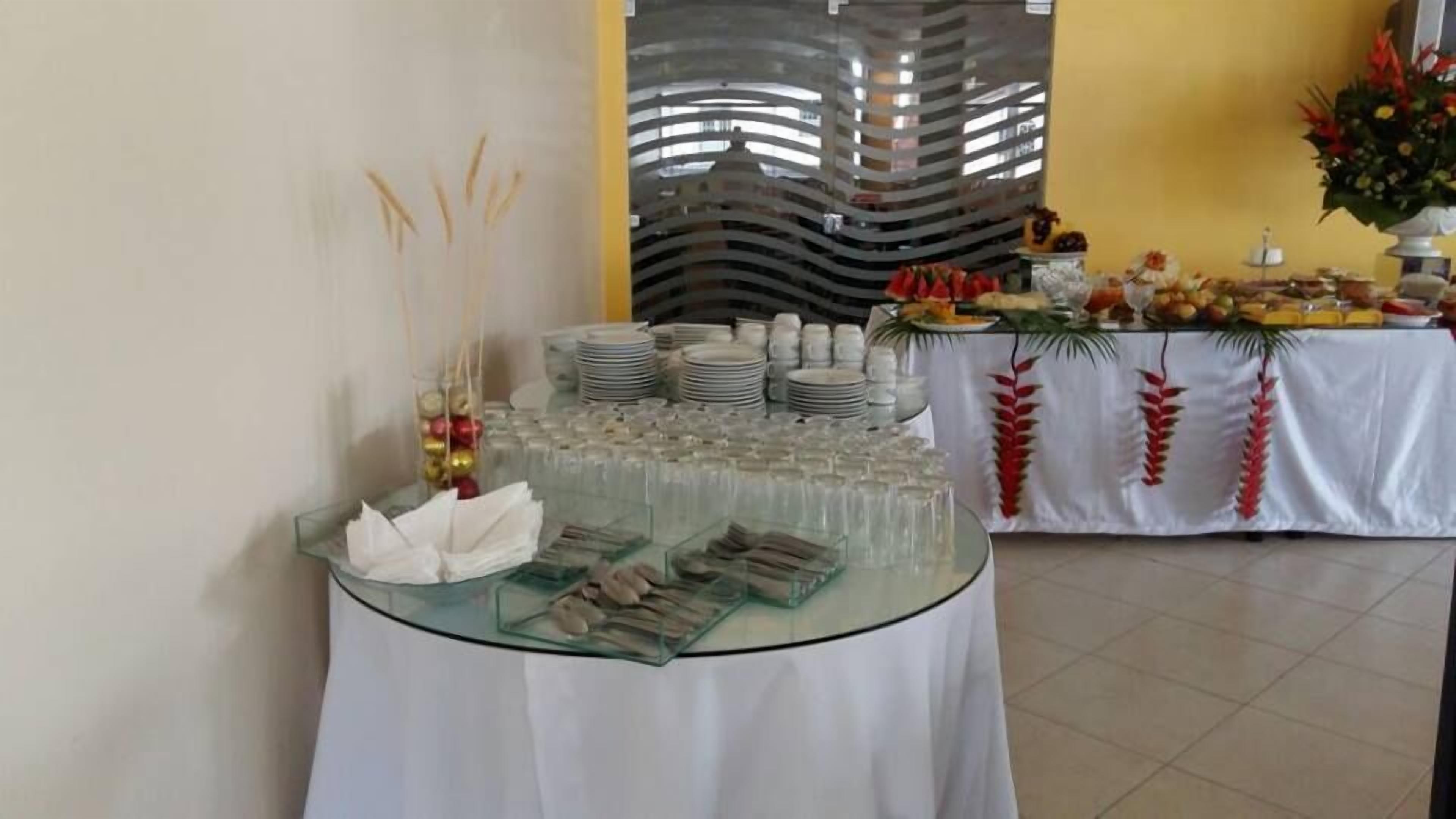 A buffet setup with glassware, plates, and napkins on a glass table, with a decorative wall and a tablecloth with palm leaves in the background.