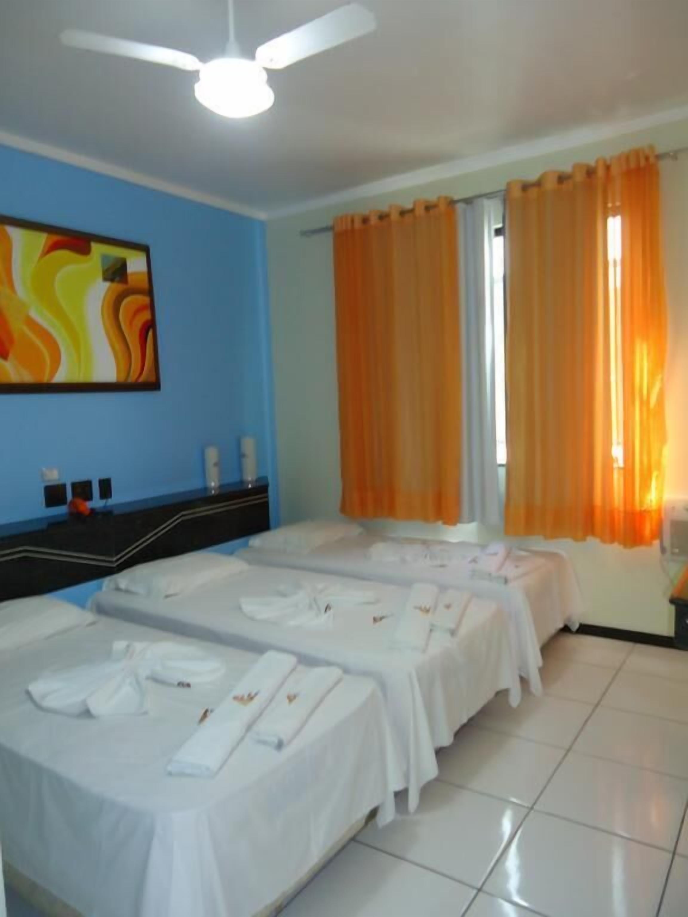 A hotel room with three beds, a ceiling fan, a painting on the wall, and orange curtains.