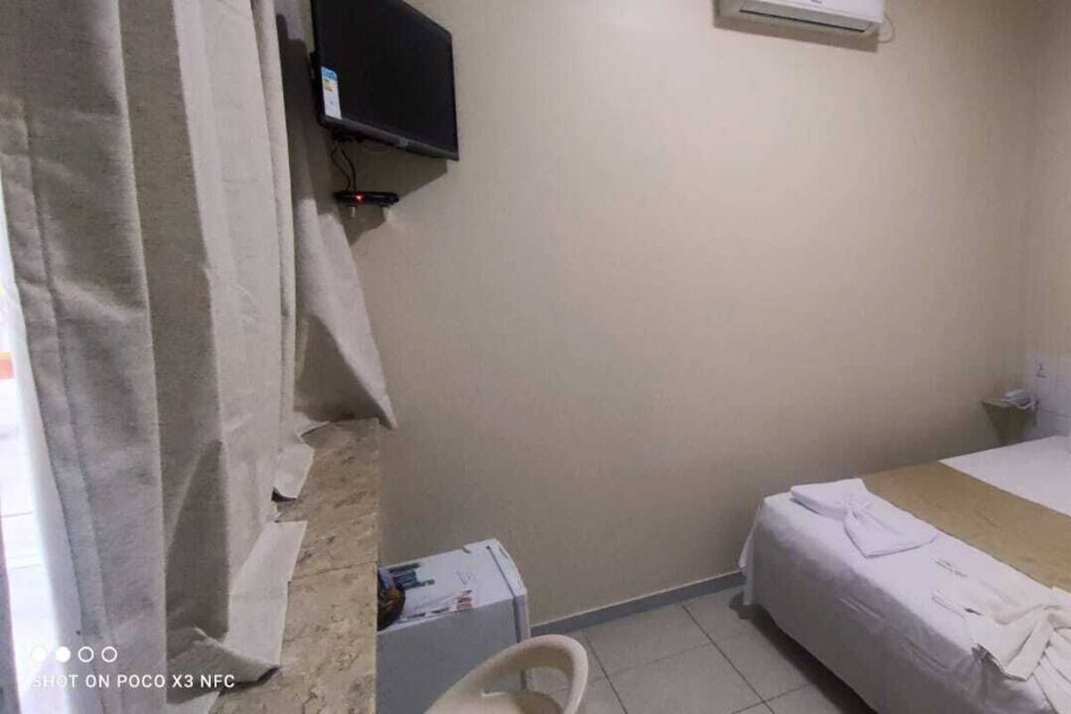 A small, simple hotel room with a bed, a nightstand, a TV mounted on the wall, and a small table with a chair.