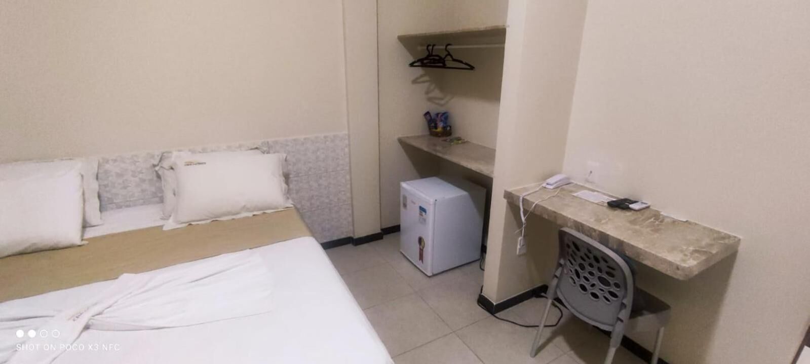 A hotel room with a bed, a small desk with a telephone, a mini-fridge, and a shelf with personal items.