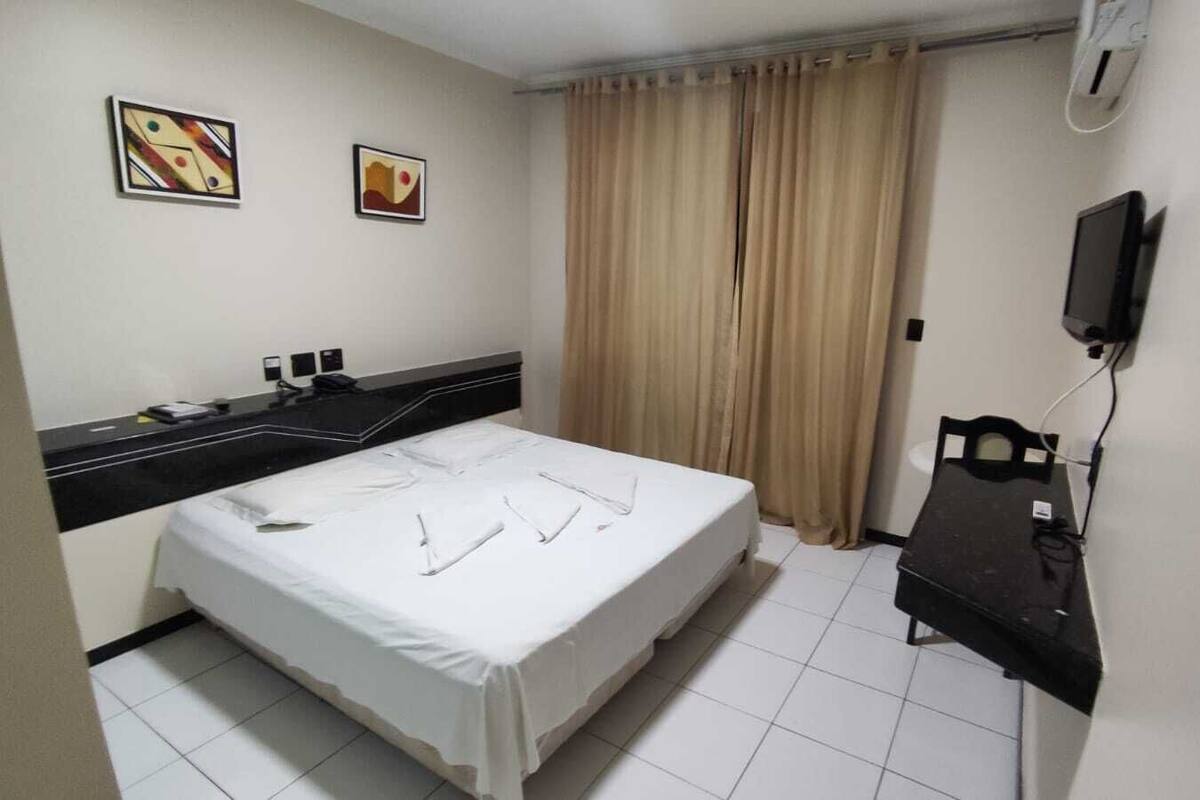 A hotel room with a bed, a desk, a chair, a TV, and two paintings on the wall.