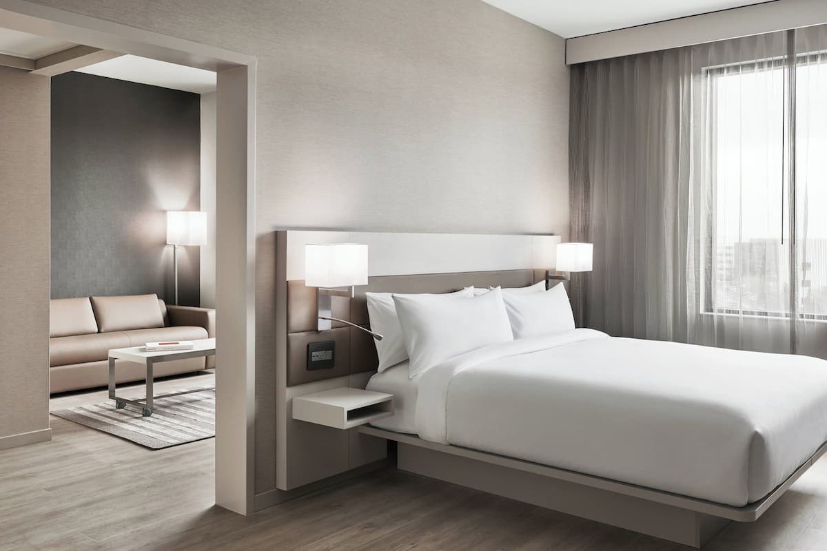 Una habitación de hotel moderna con una cama grande, mesitas de noche, un sofá y vistas de la ciudad a través de amplios ventanales.