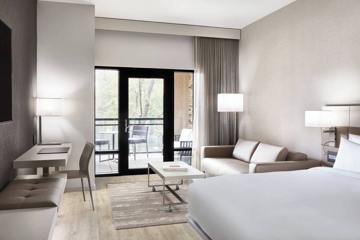 Una habitación de hotel moderna con una cama grande, un escritorio, una silla, un sofá y un balcón con vistas a árboles.