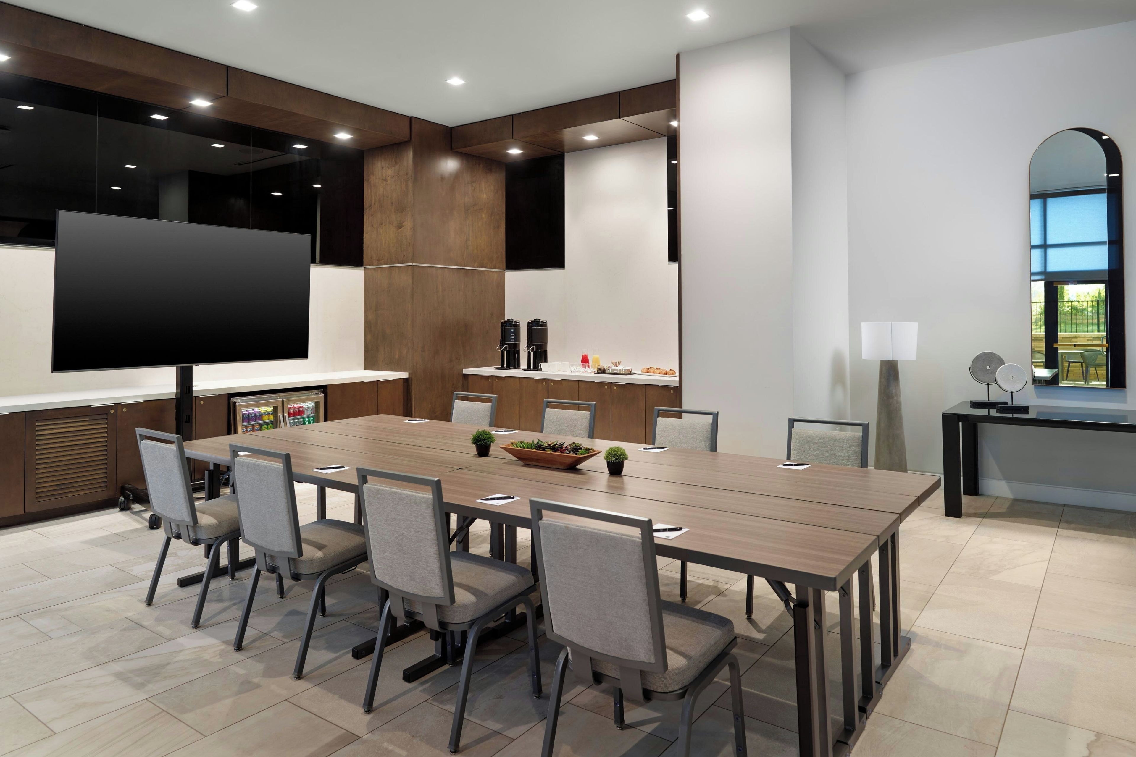 A modern conference room with a long table, chairs, a TV screen, and a coffee station.