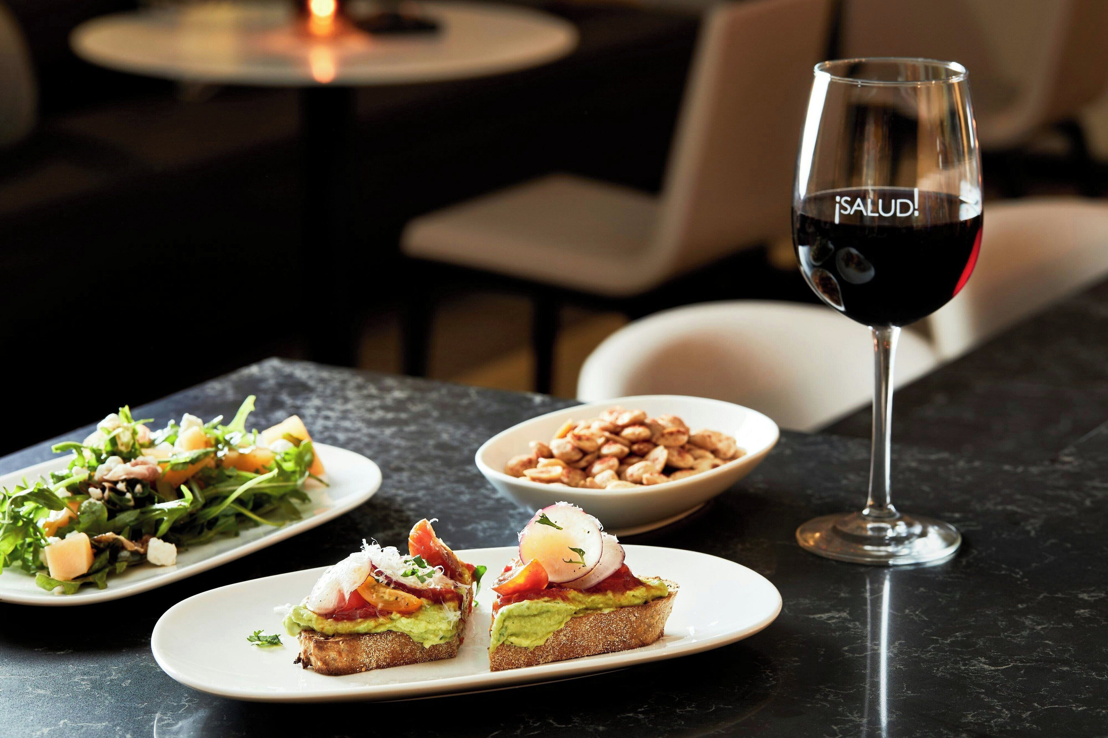 A table with a glass of red wine, two plates of appetizers, and a bowl of nuts.