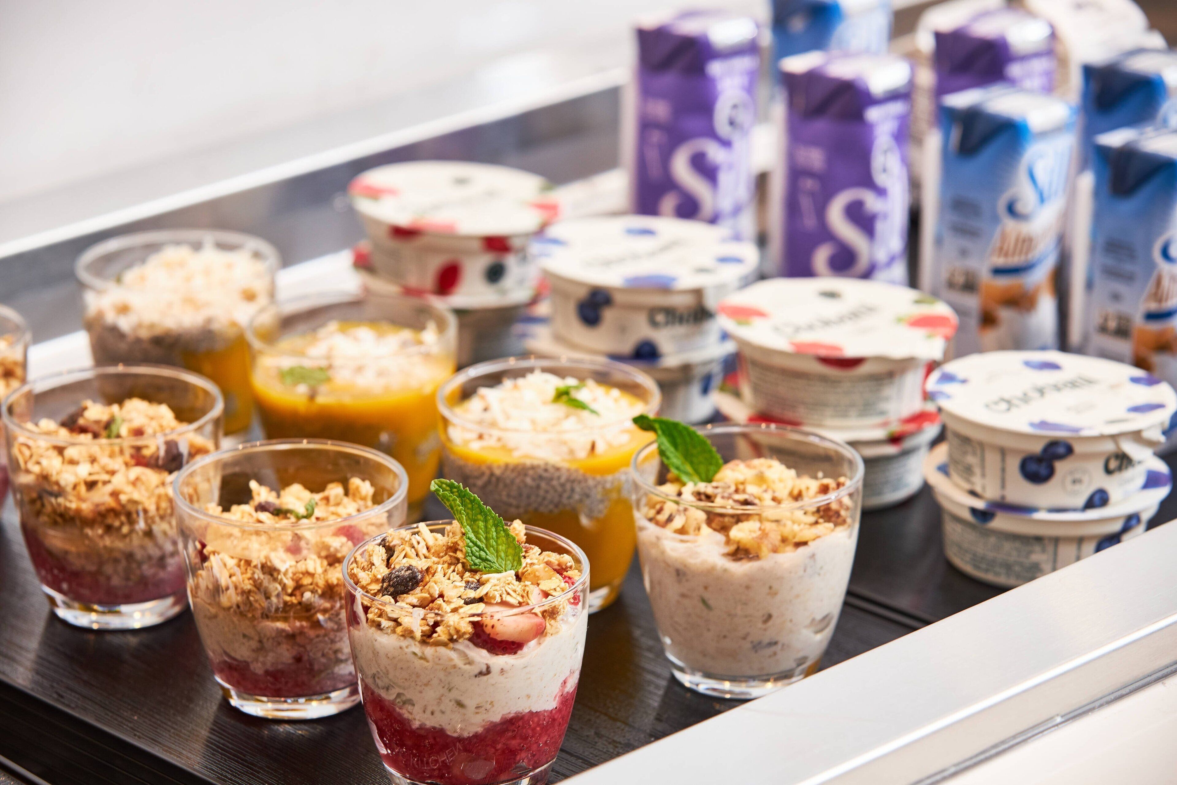 A variety of yogurt parfaits in glasses, some with granola and fruit, and several cartons of milk in the background.