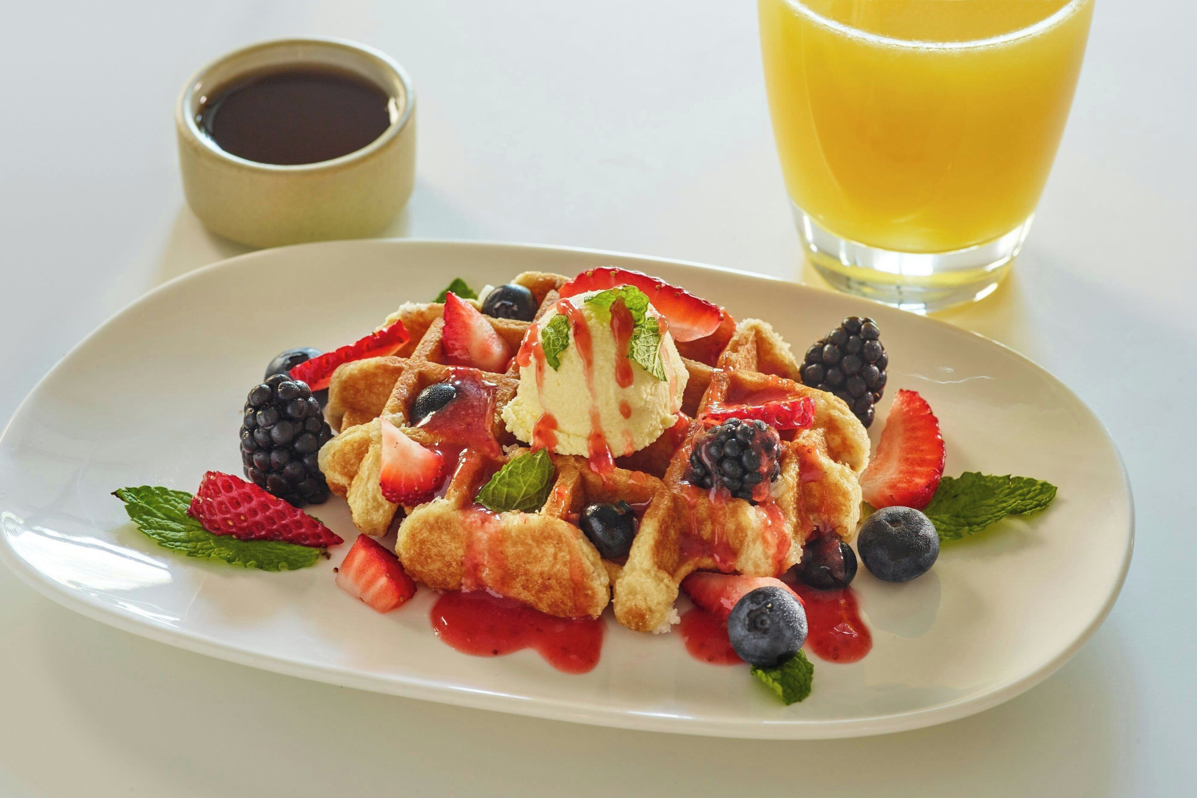 A plate of waffles with strawberries, blueberries, blackberries, and mint, accompanied by a glass of orange juice and a small cup of syrup.