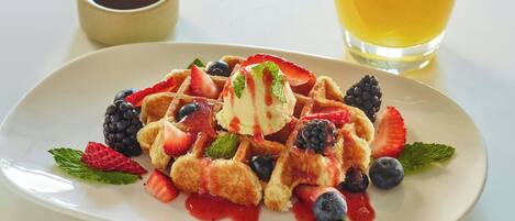 A plate of waffles with strawberries, blueberries, blackberries, and mint, accompanied by a glass of orange juice and a small cup of syrup.